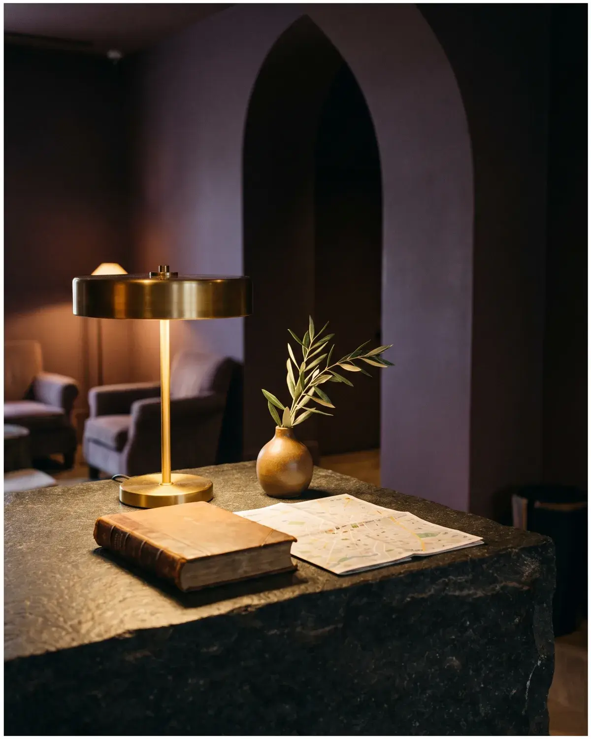 A concierge desk in a design hotel. Dark stone counter, a lit brass desk lamp, a leather-bound book, an olive stem in a small vase, a folded city map.
